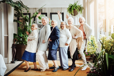 Image of six older women smiling and dancing in a conga line with one arm waving to the camera